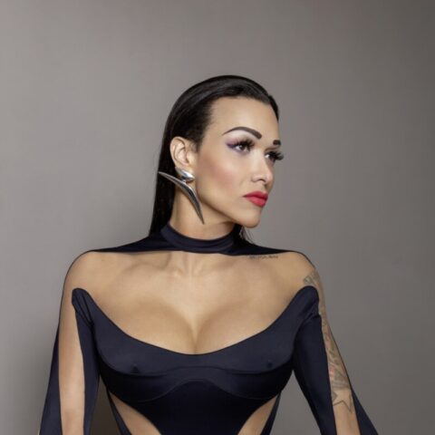 Eine Frau mit zurückgekämmtem Haar und Make-up, die einen schwarzen, futuristischen Bodysuit mit Cut-Outs trägt und vor einem grauen Hintergrund sitzt.