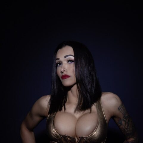Eine Frau mit dunklem Haar und einem goldenen Korsett steht vor einem dunkelblauen Hintergrund. Auf ihren Armen sind ihre Tätowierungen sichtbar.