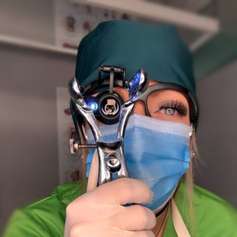 Eine medizinische Fachkraft in Kittel und Handschuhen hält ein beleuchtetes Gerät zur Augenuntersuchung an ihr eigenes Auge und schaut in die Kamera.