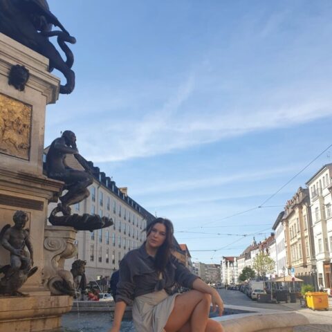 Eine Frau sitzt barfuß am Rand eines Steinbrunnens auf einem europäischen Stadtplatz. Im Hintergrund sind unter einem blauen Himmel historische Gebäude und Statuen zu sehen.
