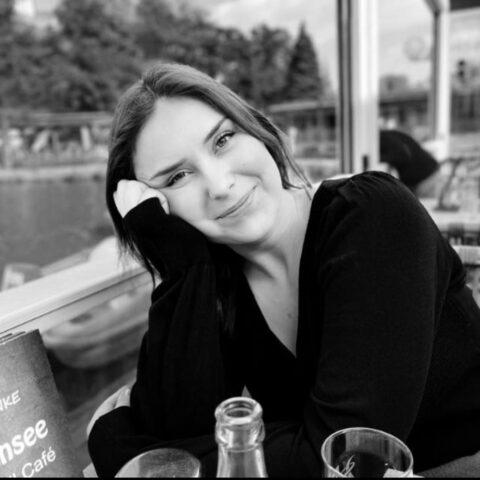 Eine schwarz gekleidete Frau mit langen Haaren sitzt an einem Cafétisch im Freien, stützt den Kopf auf die Hand und lächelt in die Kamera. Auf dem Tisch stehen mehrere Gläser und eine Speisekarte.