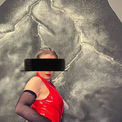 Eine Person in einem roten Lederoutfit, Netzstrümpfen und schwarzen Overknee-Stiefeln kniet vor einem strukturierten schwarz-grauen Hintergrund. Ihre Augen sind durch einen schwarzen Balken verdeckt.