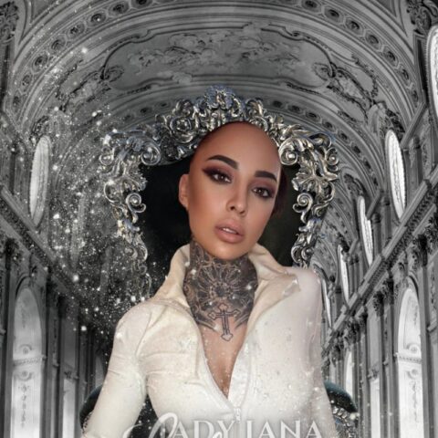 Eine Person mit einem Tattoo am Hals und einem weißen Outfit sitzt auf einem verzierten Thronsessel in einer prachtvollen architektonischen Kulisse. Der Text auf dem Bild lautet „Lady Lana Lestrange“.