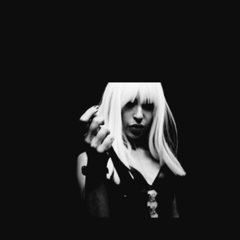 Eine Person mit langen hellen Haaren in schwarzem Outfit hält ein Mikrofon vor dunklem Hintergrund.