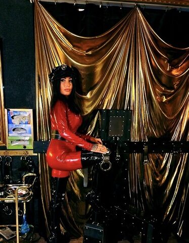 Eine Frau in einem roten Latex-Outfit und schwarzen Stiefeln posiert neben einer schwarzen mechanischen Struktur in einem Raum mit goldenen Vorhängen und Holzfußboden.