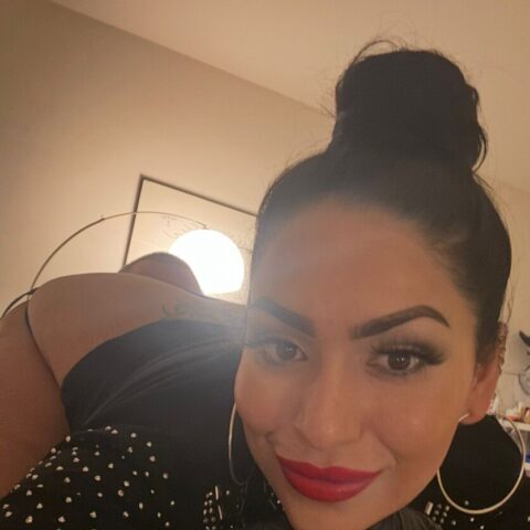 Eine Person mit dunklem Haar in einem Dutt, auffälligem Make-up, Ohrringen und schwarzen Handschuhen posiert in einem Haus und stützt ihr Kinn auf eine Hand.