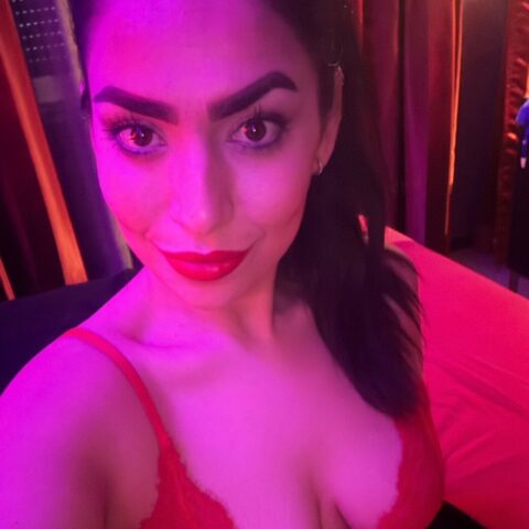 Eine Frau in einem roten Spitzenoberteil macht ein Selfie in einem Raum mit rosa und lila Licht, schaut in die Kamera und lächelt leicht.