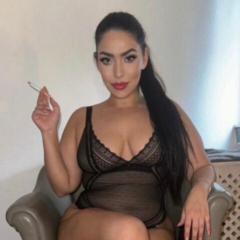 Eine Frau mit langen dunklen Haaren in einem schwarzen Netzbody sitzt im Schneidersitz in einem grauen Sessel und hat eine Zigarette in der Hand.