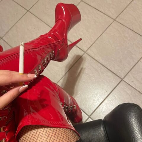 Eine Person in roten Lackstiefeln, Netzstrümpfen und Spitzenstrumpfband sitzt auf einem schwarzen Stuhl und hält eine angezündete Zigarette über einen gefliesten Boden.