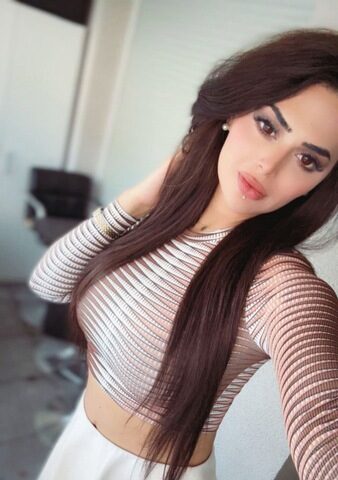 Eine Frau mit langen dunklen Haaren macht ein Selfie. Sie trägt ein gestreiftes, langärmeliges Oberteil und einen weißen Rock und steht in einem Haus in der Nähe eines Stuhls und eines Tisches.