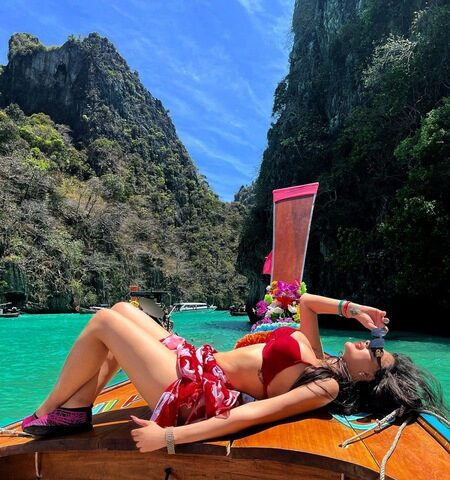 Eine Frau im roten Bikini entspannt sich auf dem Bug eines Holzbootes in türkisfarbenem Wasser, umgeben von felsigen Klippen und üppigem Grün unter einem strahlend blauen Himmel.