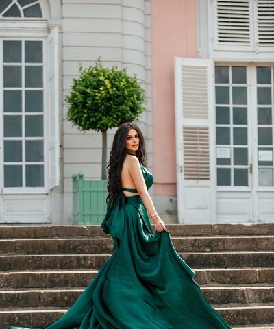 Eine Frau in einem langen, fließenden grünen Kleid steht auf einer Steinstufe im Freien vor einem Gebäude mit großen Fenstern und weißen Fensterläden.