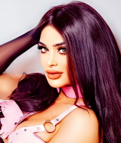 Eine Frau mit langen dunklen Haaren trägt ein rosafarbenes Halsband und ein rosafarbenes Geschirr mit flügelförmigen Details und schaut direkt in die Kamera.