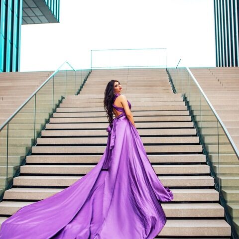 Eine Frau in einem fließenden violetten Kleid steht auf einer breiten Freitreppe und blickt über ihre Schulter, während im Hintergrund moderne Glasgebäude zu sehen sind.