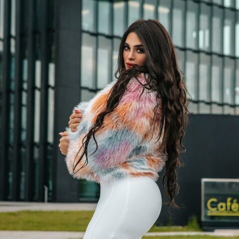 Frau mit langen dunklen Haaren, die eine bunte Kunstpelzjacke und einen weißen Rock trägt, steht im Freien vor einem modernen Glasgebäude.