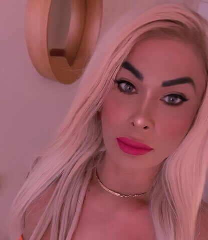 Person mit langen blonden Haaren und auffälligem Make-up, die ein farbenfrohes trägerloses Blumentop und eine goldene Halskette trägt und eine Tätowierung auf einem Arm zeigt, steht in einem Haus.